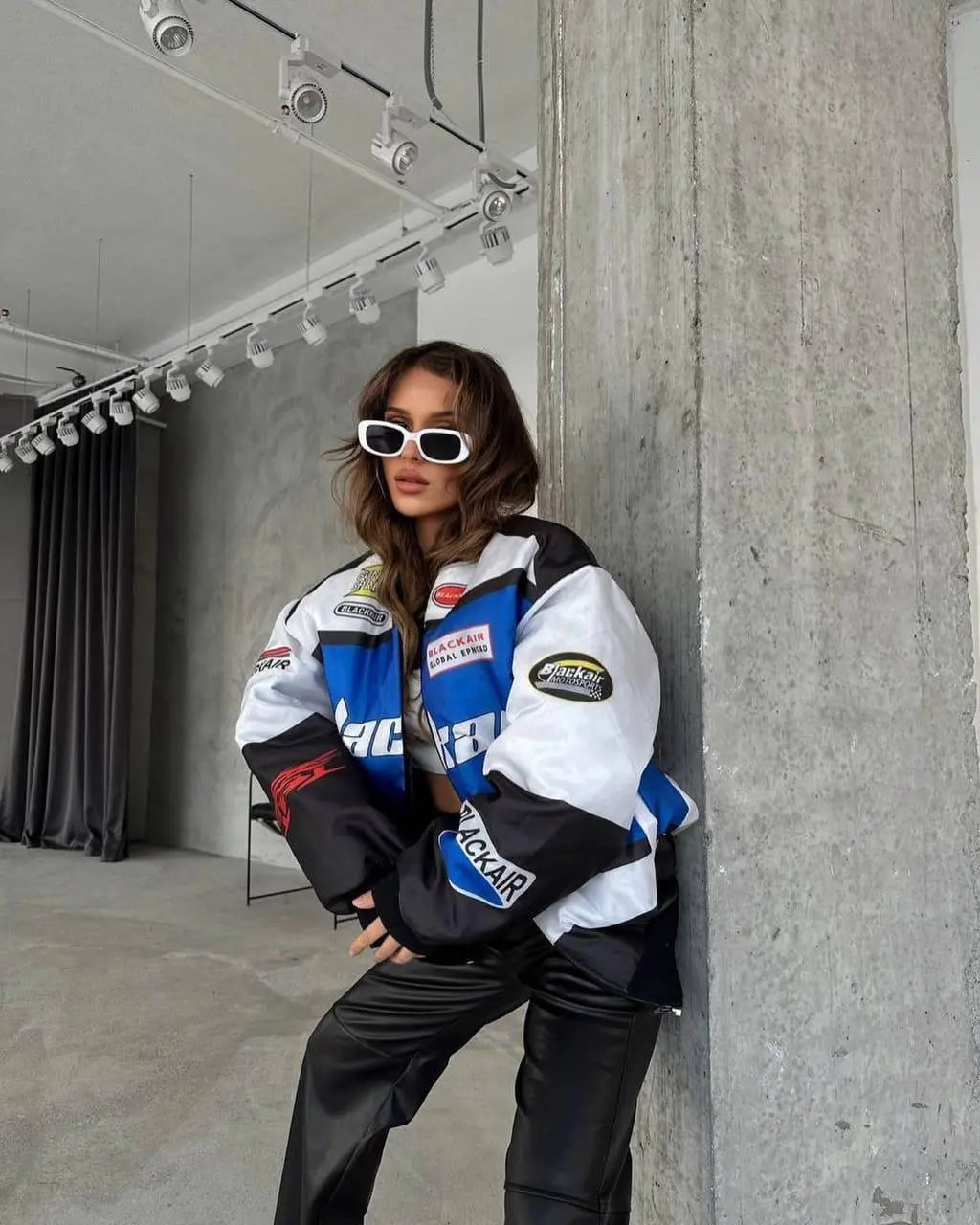 Woman in streetwear style, wearing a blue and white racing jacket, black pants, and white sunglasses, posing indoors against a concrete wall.