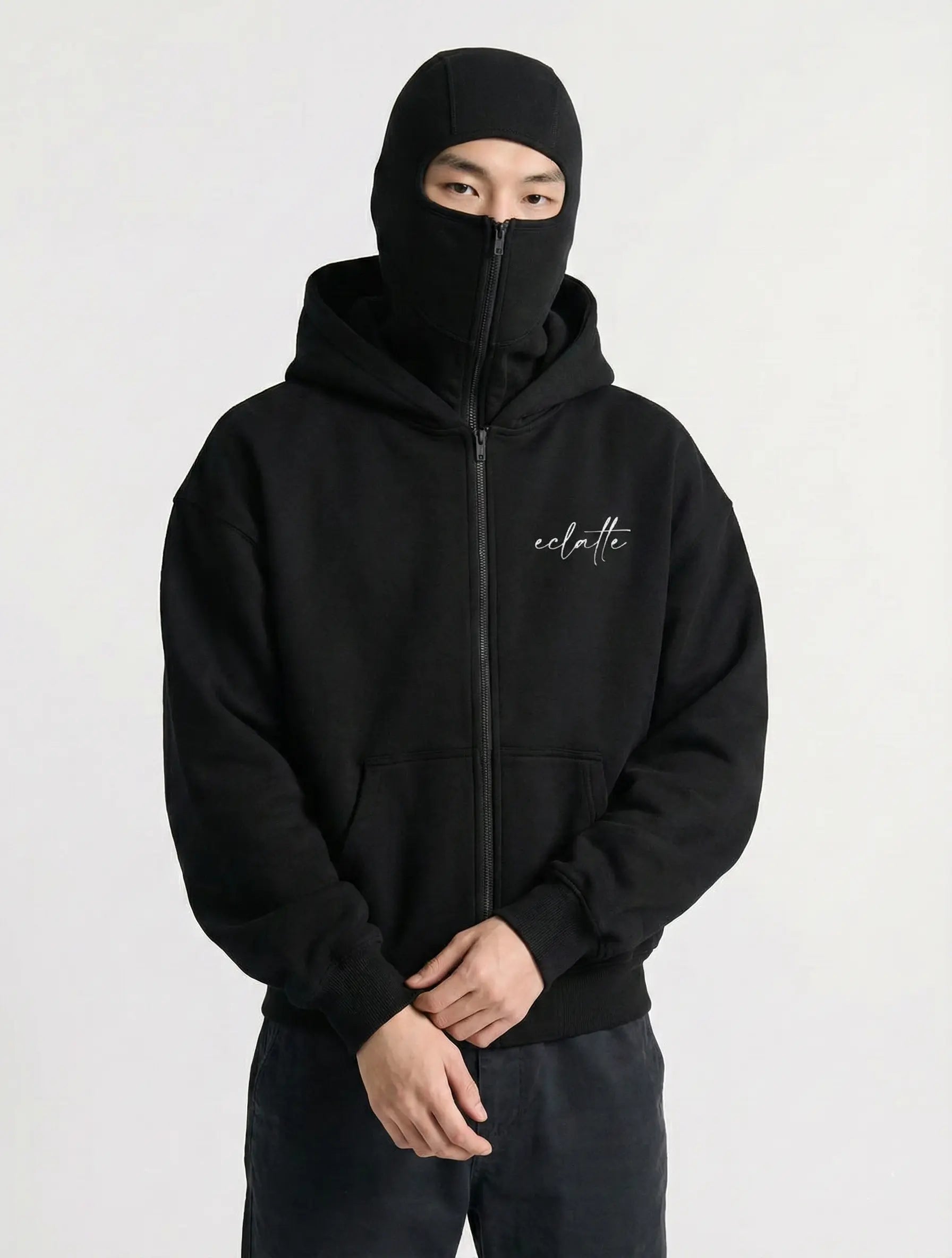 Man in black zip-up hoodie with face cover, minimalist streetwear style, white script logo
