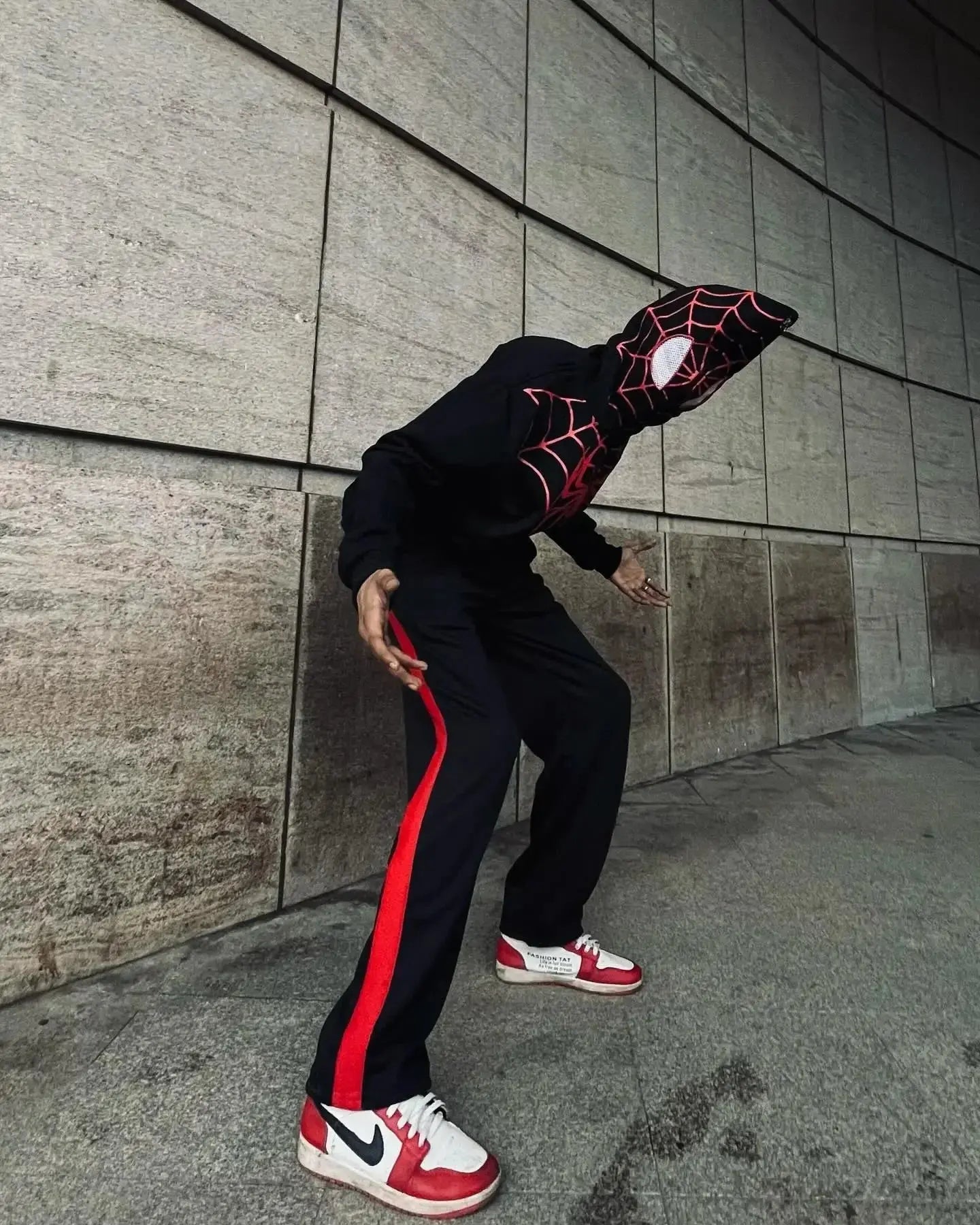 Person wearing a black Spider-Man zip-up hoodie with web design, matching pants, and red sneakers.